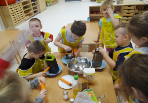 Igorek, Lenka i Antoś ścierają marchewkę na tartce - pozostałe dzieci obserwują.